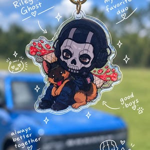 May include: A clear acrylic keychain featuring a chibi-style character wearing a skull mask and a German Shepherd dog. The design includes text reading Riley & Ghost, my favourite duo, good boys, and always better together, with small star and paw icons.