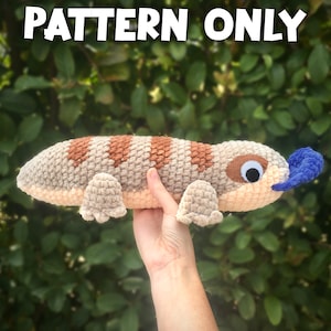 May include: Crochet pattern for a plush lizard toy. The lizard is brown and beige with a blue tongue. The toy is being held in a hand.