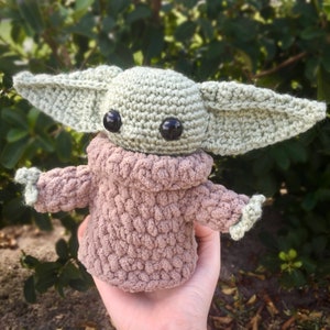 Puede incluir: Mu&ntilde;eco de ganchillo de un personaje con orejas grandes y ojos negros. El mu&ntilde;eco es de color verde claro con una t&uacute;nica marr&oacute;n y brazos peque&ntilde;os. El mu&ntilde;eco est&aacute; hecho a mano y parece un personaje de una popular serie de ciencia ficci&oacute;n.