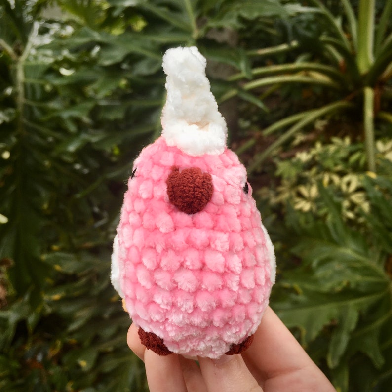 Op de afbeelding: Een roze gehaakte vogel met een witte gehaakte kroon en een rood hart op zijn borst.
