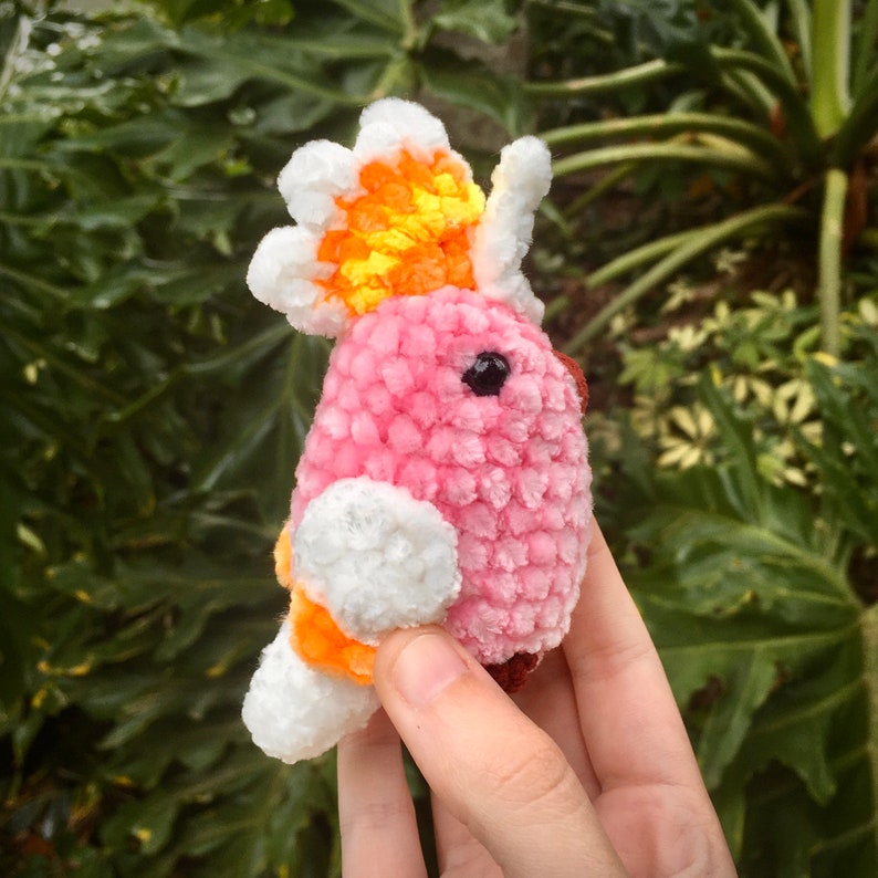 Op de afbeelding: Een roze gehaakte vogel met een witte en oranje kuif. De vogel is gemaakt van zacht, pluche garen en heeft een zwart oog.