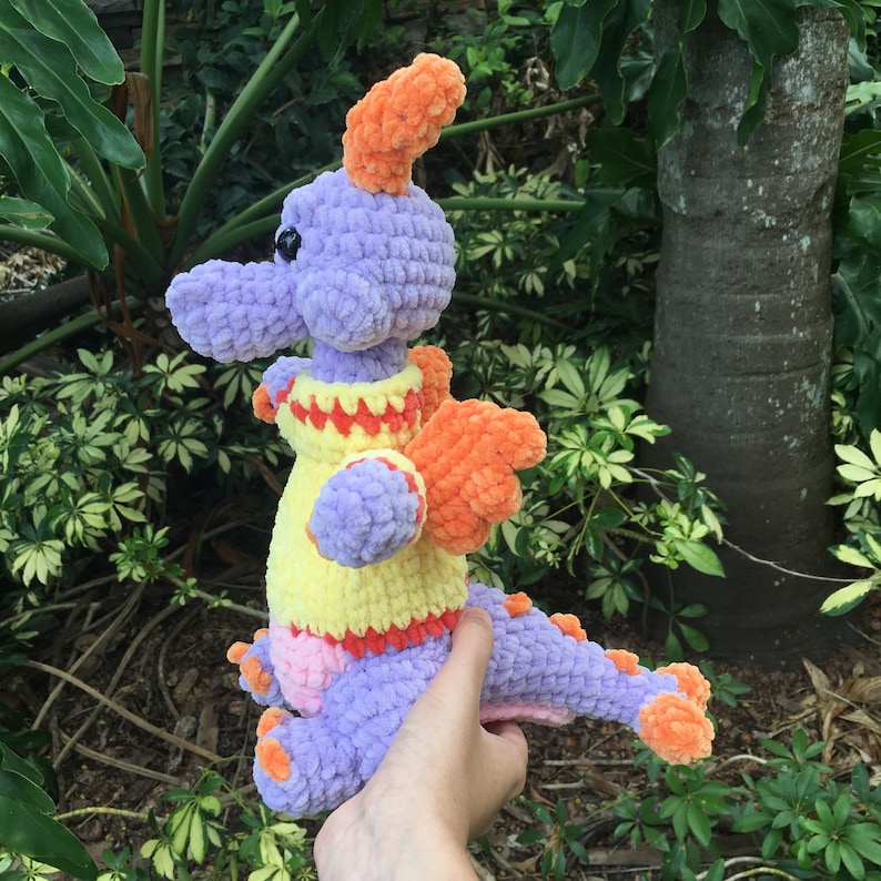 Puede incluir: Un juguete de peluche de drag&oacute;n de crochet morado y naranja con un su&eacute;ter amarillo y rojo. El drag&oacute;n tiene una cola larga y alas naranjas.