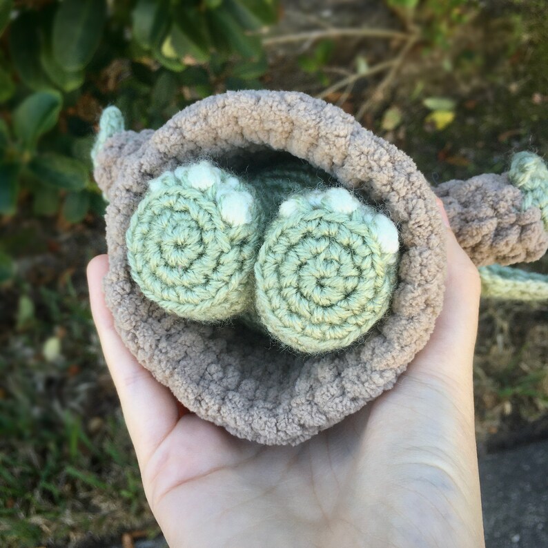 Puede incluir: Una mu&ntilde;eca de ganchillo hecha a mano con pies verde claro y cuerpo marr&oacute;n. Los pies de la mu&ntilde;eca son redondos con detalles blancos. La mu&ntilde;eca se sostiene en una mano, con un fondo verde frondoso.