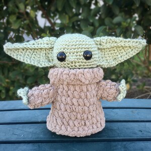 Puede incluir: Mu&ntilde;eco de ganchillo de un personaje con orejas grandes, ojos negros y una t&uacute;nica marr&oacute;n. El mu&ntilde;eco es de color verde claro y marr&oacute;n, con un aspecto texturizado. El mu&ntilde;eco est&aacute; hecho a mano y parece ser un personaje de una popular franquicia de ciencia ficci&oacute;n.