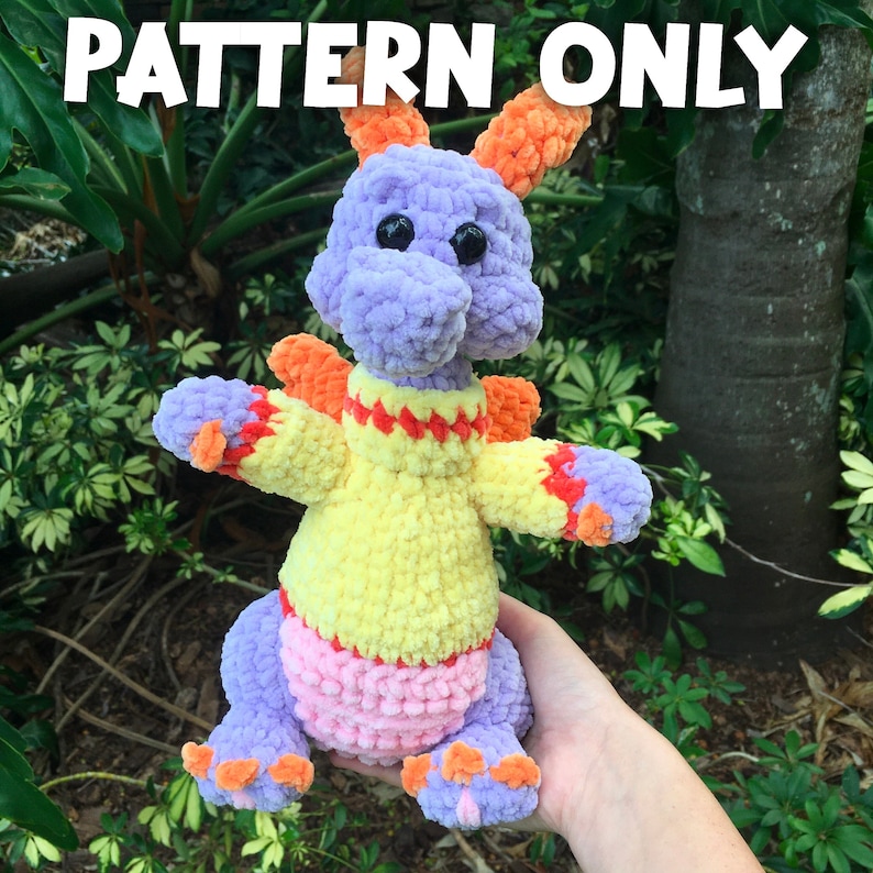 Puede incluir: Un drag&oacute;n de peluche tejido a crochet con hilo morado, naranja, amarillo y rosa. El drag&oacute;n lleva un su&eacute;ter amarillo con detalles naranjas y rojos.