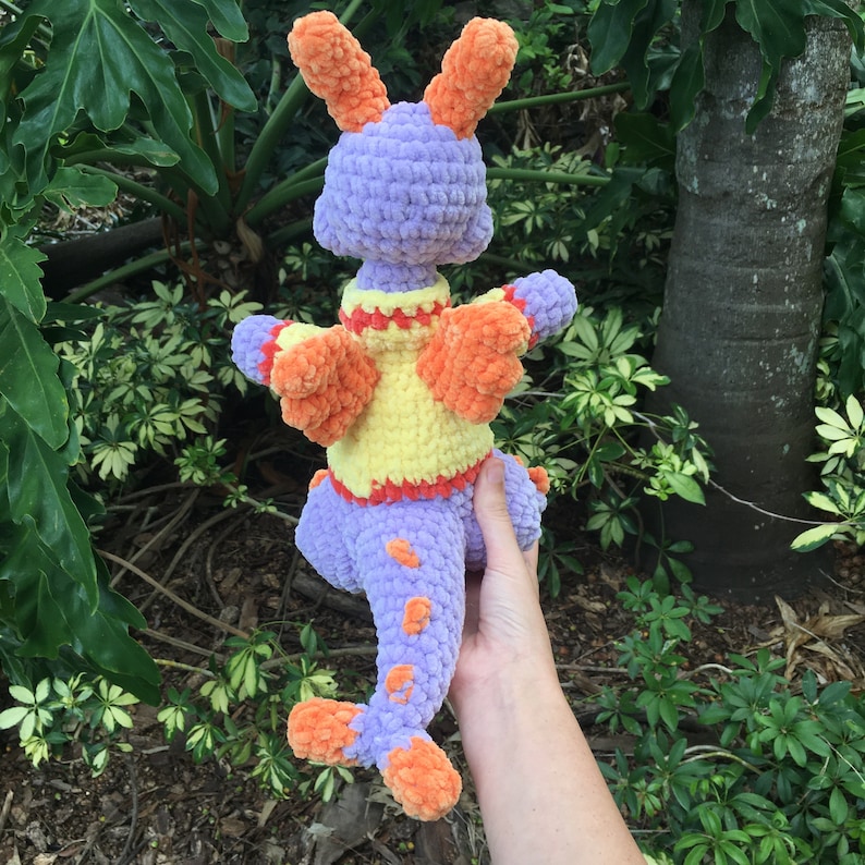 Puede incluir: Un juguete de drag&oacute;n de crochet morado y naranja con detalles amarillos y naranjas. El drag&oacute;n tiene una cola larga y alas grandes.