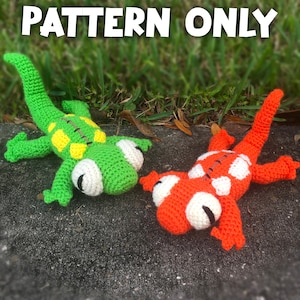 May include: Two crocheted lizard toys, one green with yellow spots and one orange with white spots. Both have black eyes and are lying on a grey surface.