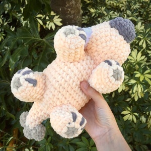 May include: A crocheted plush toy in the shape of a pink pig with gray feet and a gray snout. The pig is lying on its back with its legs in the air.