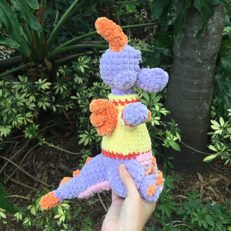 Puede incluir: Un drag&oacute;n de crochet morado con detalles en naranja y amarillo. El drag&oacute;n est&aacute; sosteniendo sus alas extendidas y tiene una expresi&oacute;n amigable.