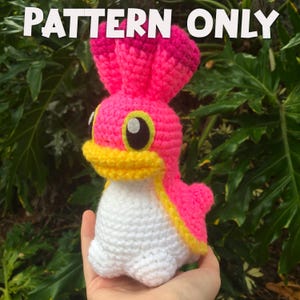 May include: A handmade crochet figure in bright pink, yellow, and white, with the text "PATTERN ONLY" at the top. The figure has large black and yellow eyes and a yellow beak. The crochet animal is held in front of a green leafy background.