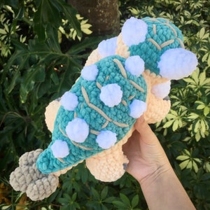 May include: A crocheted plush toy shaped like an ankylosaurus. The toy is teal green with white spikes and a light peach belly.