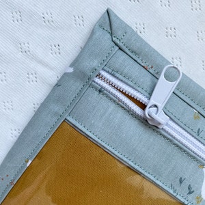Könnte beinhalten: Eine hellblaue Reißverschlusstasche mit einem transparenten Plastikfenster und einem weißen Reißverschluss. Die Tasche hat ein Blumenmuster und ein senfgelbes Futter.