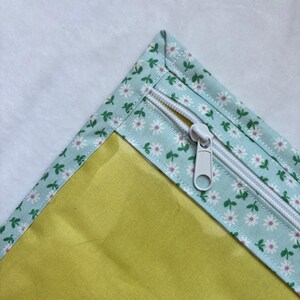 Può includere: Una custodia in tessuto con un pannello giallo senape a tinta unita e un bordo azzurro decorato con piccole margherite bianche e foglie verdi. La custodia presenta una chiusura a zip bianca con un cursore in plastica bianca.