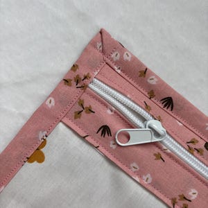 Può includere: Primo piano di un tessuto rosa e bianco con cerniera. Il tessuto rosa presenta un motivo floreale con piccoli fiori marroni e bianchi. La cerniera bianca è aperta, rivelando l'interno del tessuto.