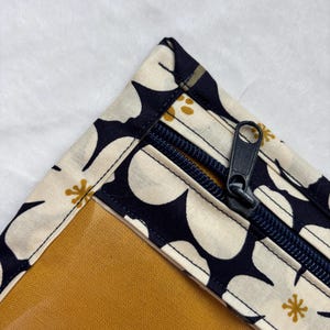 Op de afbeelding: Close-up van een bloemmotief etui met een marineblauwe rits. De stof heeft een crèmekleurige achtergrond met grote witte bloemvormen en kleine gouden accenten. De binnenkant is mosterdgeel.