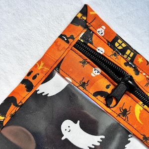 Op de afbeelding: Een tasje met rits en een oranje stoffen rand met een patroon van zwarte spinnen, witte schedels en gele maansikkels. Het hoofdvak is gemaakt van transparant materiaal bedrukt met witte spookfiguren.