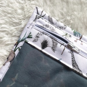 Może przedstawiać: Biała kosmetyczka z przezroczystym okienkiem i nadrukiem zwierząt z safari. Nadruk przedstawia żyrafy, palmy i inne rośliny w odcieniach szarości i zieleni. Zamek błyskawiczny jest biały.