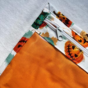 Può includere: Una pochette in tessuto con bordo bianco e una stampa colorata di zucche e foglie autunnali. La pochette presenta una chiusura a zip bianca con un cursore in metallo argentato e una fodera interna arancione tinta unita visibile.