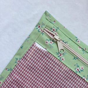 Op de afbeelding: Een stoffen etui met een lichtgroene rand voorzien van een klein roze en wit bloemenpatroon. Het hoofdvlak van het etui bestaat uit rood-witte vichy-ruitjesstof. Een beige rits met een bijpassende plastic ritstrekker is aan de bovenkant genaaid.