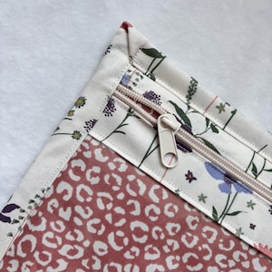 Op de afbeelding: Close-up van een etui met ritssluiting met een bloemenprintrand en een rood-witte luipaardprint binnenkant. De rits is beige en het etui staat op een witte achtergrond. De bloemenprint bevat paarse, blauwe en groene bloemen.