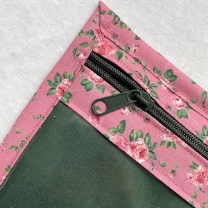 Può includere: Un angolo di una pochette con cerniera con un motivo floreale rosa. Il tessuto presenta un motivo ripetuto di rose rosa e foglie verdi su sfondo rosa. La cerniera è nera con un tirante verde scuro.