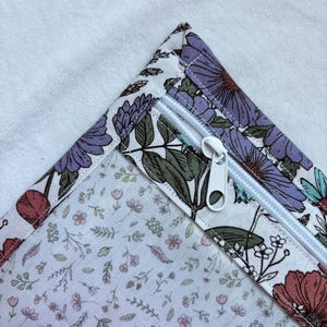 Può includere: Una custodia in tessuto con sfondo bianco e una stampa floreale in scala ridotta nei toni del rosa, verde e marrone. Il bordo superiore presenta una finitura a contrasto con fiori grandi viola e rossi e foglie verdi, chiusa da una cerniera in plastica bianca.