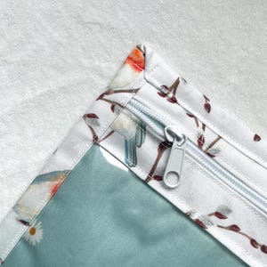 Può includere: Una custodia in tessuto con sfondo bianco e motivo di piccoli uccelli e rami, arricchita da una sezione color verde acqua con motivi a margherite bianche. La custodia presenta una chiusura a zip bianca con cursore in plastica abbinato.