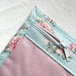 Op de afbeelding: Close-up van een stoffen etui met een bloemen- en stippenpatroon. De etui heeft een lichtblauwe rand met witte stippen en een bloemenpatroon. De binnenkant is roze met witte stippen en heeft een zilveren rits.