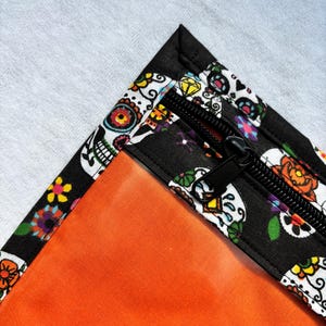 Può includere: Una custodia in tessuto nero con un motivo colorato di teschi di zucchero e fiori, dotata di una chiusura a zip nera. La fodera interna è in tessuto arancione brillante a tinta unita. Il design presenta teschi bianchi con fiori.