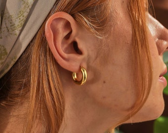 May include: A close-up of a person's ear wearing a pair of gold hoop earrings. The earrings are thick and circular. The person has red hair and is wearing a light-colored scarf with a green floral pattern. The background is blurred.