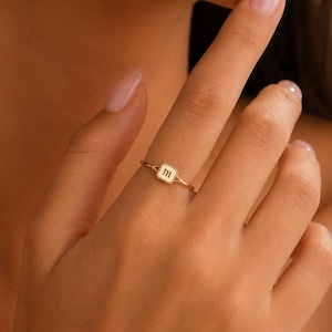 May include: A gold-toned ring featuring a small, square-shaped centerpiece with a textured border and the lowercase letter m engraved in the center. The band is thin and metallic, encircling the finger with a polished finish and simple design.