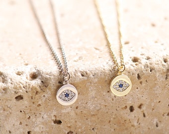 May include: Two delicate necklaces with a small evil eye pendant. The pendant is made of silver and gold and features a blue stone in the center. The necklaces are on a light brown textured surface.