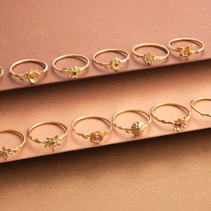May include: A collection of 14 gold rings with floral designs and colorful gemstones. The rings are arranged in two rows on a brown surface.