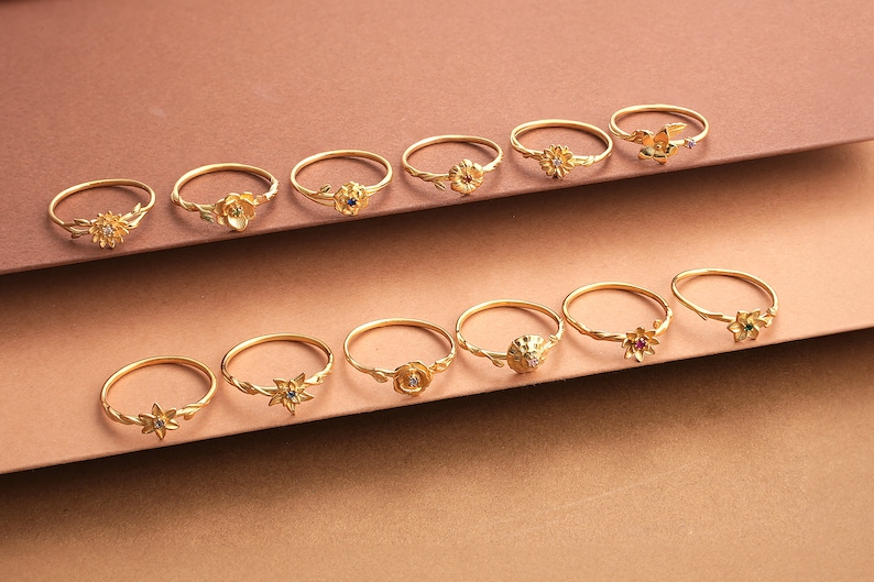 May include: A collection of 14 gold rings with floral designs and colorful gemstones. The rings are arranged in two rows on a brown surface.