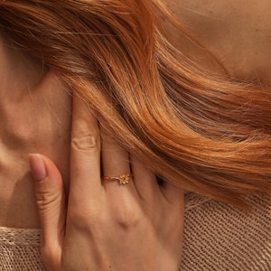 May include: A gold ring with a small flower design. The ring is worn on the finger of a person with long, red hair.