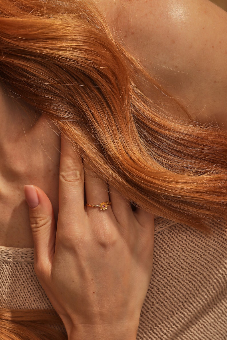 May include: A gold ring with a small flower design. The ring is worn on the finger of a person with long, red hair.