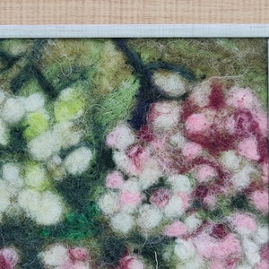May include: A framed felt artwork depicting a close-up of a floral arrangement. The piece features white, pink, and burgundy flowers with green foliage. The frame is a light blue-grey colour, and the background is light wood.