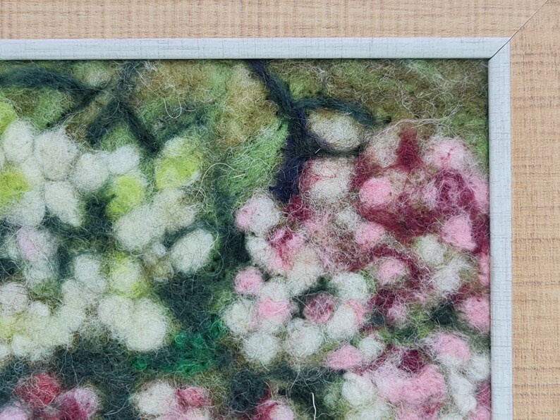 May include: A framed felt artwork depicting a close-up of a floral arrangement. The piece features white, pink, and burgundy flowers with green foliage. The frame is a light blue-grey colour, and the background is light wood.