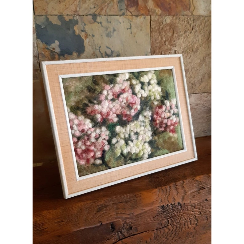May include: A framed floral artwork with pink and white flowers. The frame is light-coloured with a white inner border. The artwork is displayed on a wooden surface. The piece is reminiscent of a needlepoint design.