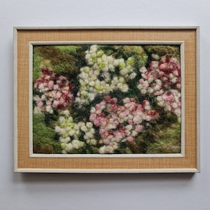 May include: A framed textile artwork depicting a close-up of a floral design. The piece shows clusters of white and pink flowers against a green backdrop. The frame has a light brown textured border and a white inner edge.