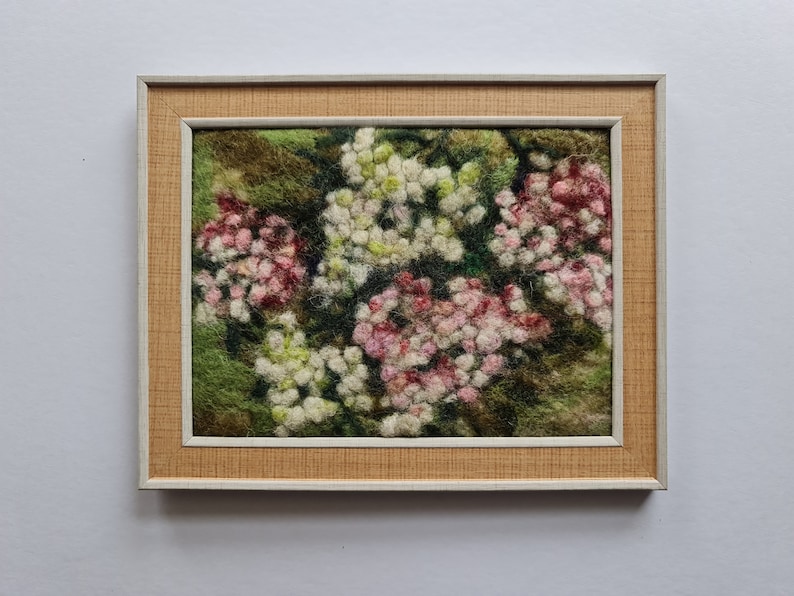 May include: A framed textile artwork depicting a close-up of a floral design. The piece shows clusters of white and pink flowers against a green backdrop. The frame has a light brown textured border and a white inner edge.