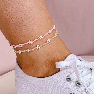 Könnte beinhalten: Zwei zarte Fußkettchen mit kleinen weißen Blumenperlen auf einer rosa und silbernen Schnur. Die Fußkettchen werden am Knöchel einer Person über einem weißen Canvas-Schuh mit dem Wort "SUPERGA" an der Seite getragen.