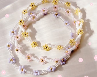 May include: Three layered beaded bracelets with small flower charms in shades of purple, pink, and yellow. The bracelets are made with clear beads and are layered on top of each other.