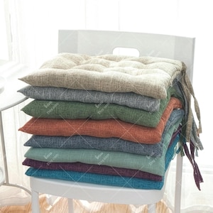 Puede incluir: Una pila de cojines de asiento con mechones en varios colores, incluyendo beige, gris, verde, naranja, azul, morado y turquesa. Los cojines están hechos de una tela texturizada y están atados con lazos de tela. Están apilados en una silla blanca.