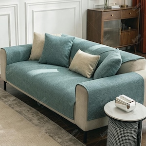May include: A modern sofa with a teal cover and several throw pillows in beige and teal. The sofa has a light beige frame and black legs. A small side table with a marble top and a patterned stool are in front of the sofa.