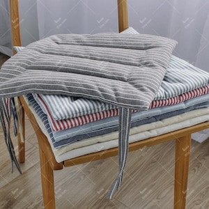 May include: A wooden chair supports a stack of striped chair cushions in shades of grey, red, and blue. The top cushion is grey with white stripes and has tie fastenings. The cushions are made of a soft fabric, suitable for indoor use.