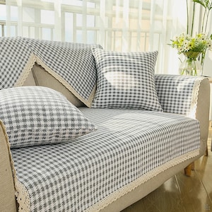 Puede incluir: Un sofá con funda a cuadros grises y blancos y cojines a juego. La funda tiene un ribete de encaje. El sofá está frente a una ventana con cortinas blancas. Un jarrón con flores está sobre una mesa auxiliar.