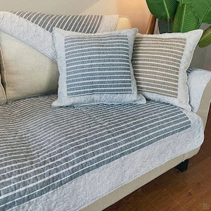 May include: A gray and white striped sofa cover and two matching square throw pillows. The pillows and cover have a quilted design. The sofa is beige with dark legs. The pillows are gray and brown.
