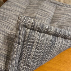 May include: A rectangular cushion featuring a textured fabric with a pattern of thin, vertical dark blue and white stripes. The cushion has a thick, piped edge detail and a soft, padded construction with visible stitching along the seams.