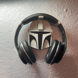 May include: A black and silver headset rests on a wall-mounted holder shaped like a Mandalorian helmet. The holder is gray with black details. The wall has a textured, gray finish.
